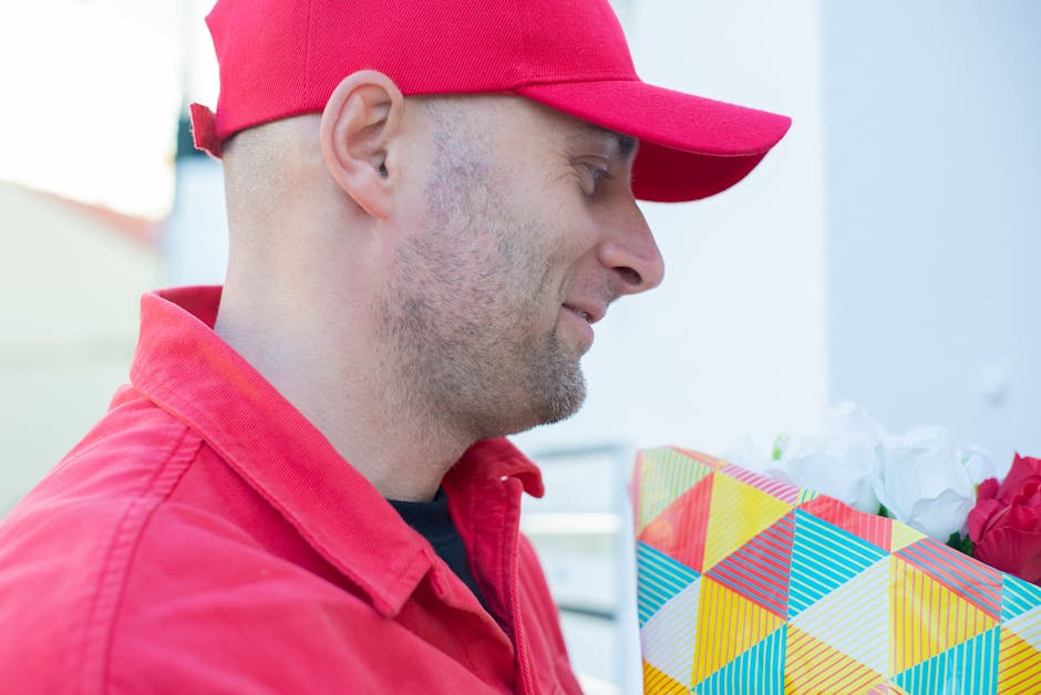 A delivery person wearing a bright red cap and matching red jacket holds a floral gift, wrapping a bouquet of mixed flowers in colorful patterned paper, including red and white blooms, potentially ros