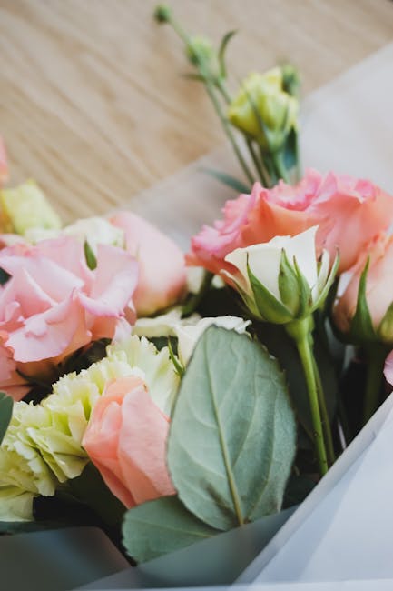 Close-up of a delicate floral arrangement featuring soft pink and cream lisianthus flowers with ruffled petals, accompanied by fresh green leaves and buds. The bouquet is partially wrapped in transluc