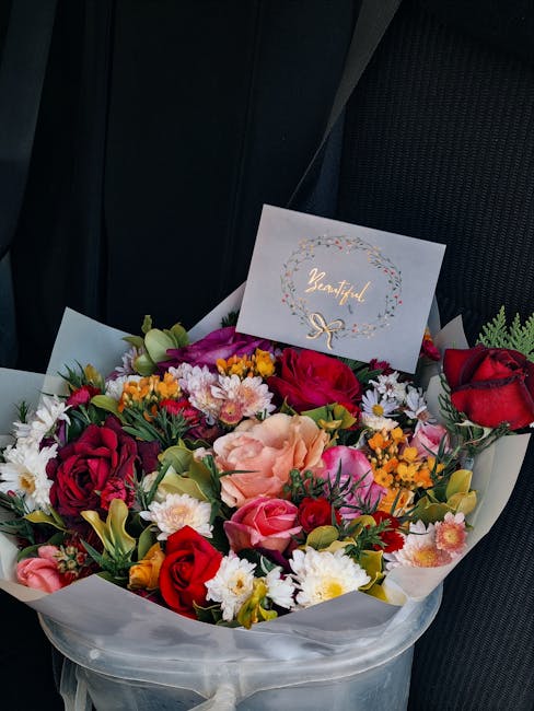 A vibrant floral arrangement featuring a variety of fresh flowers, including deep red roses, soft pink carnations, white daisies, and small yellow and orange blooms, all arranged in a round bouquet wr