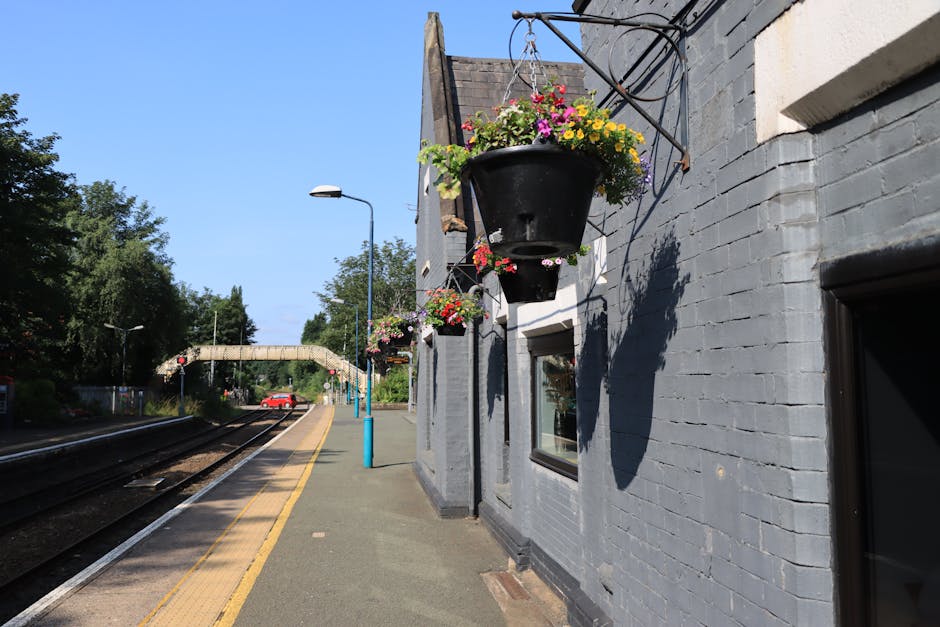 A series of black hanging flower baskets attached to a grey brick wall outside Oxted Station, RH8, filled with colorful flowers including pink, yellow, and purple blooms. The baskets are suspended by 