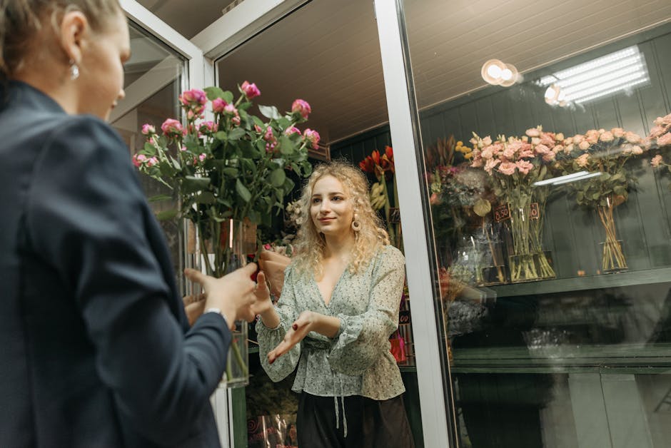 A young woman with curly blonde hair and light skin is standing inside a flower shop, receiving a bouquet of pink roses from a florist dressed in dark clothing. The bouquet consists of fresh, vibrant 