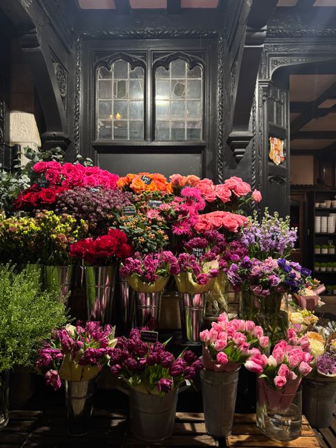 An indoor display of fresh floral arrangements featuring a variety of flowers in vibrant pinks, purples, oranges, and reds, arranged in metallic vases placed on a dark wooden surface. The flowers incl