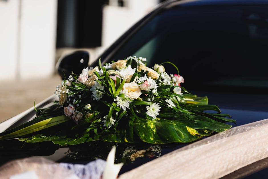 A floral arrangement featuring pale cream roses, white daisies, and small pink buds, accented with delicate sprigs of baby's breath and lush green foliage, resting on the hood of a black car. The bouq