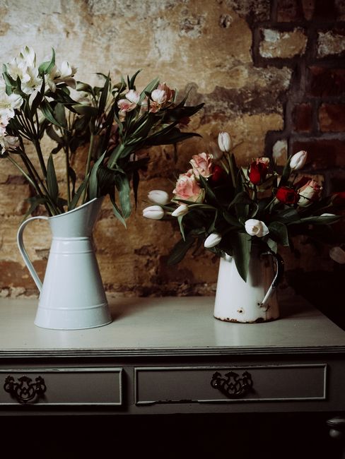 Two floral arrangements in vintage-style white metal jugs sit on a grey wooden sideboard against an exposed brick wall. The left jug contains a bouquet of white tulips with long green stems and leaves